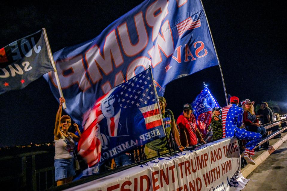 Supporters carrying 8-foot-tall Trump flags gather, with one person wearing a jumpsuit that is half red and white stripes and half white stars on a blue background, clambers over a barrier covered with a banner that says (in part): President Trump and Those ... [not visible] Stand With Him.