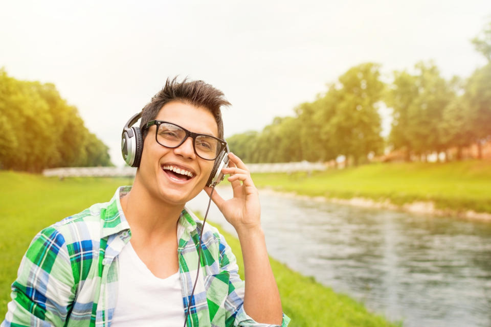 Young male adult outdoors walking in the green grass next to a stream with headphones and a big smile.