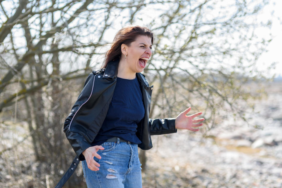 A woman stands outdoors, screaming with a distressed expression, wearing a black jacket and ripped jeans. Sparse trees and rocks are in the background