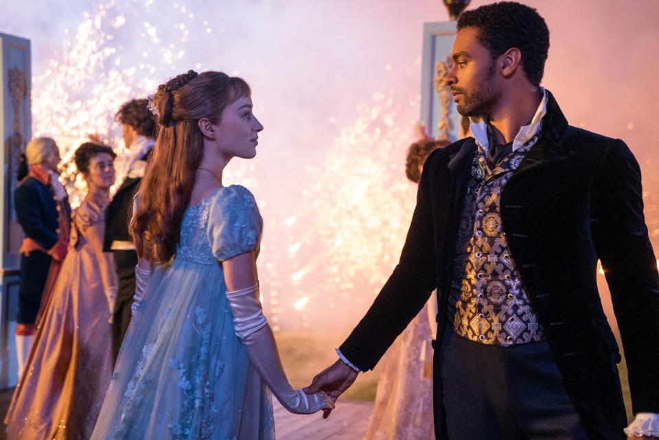 daphne and simon dancing in bridgerton. daphne is wearing a sheer blue gown, her hair worn half up, and simon is wearing a shining waistcoast and black overcoat. they are looking intently into each others eyes