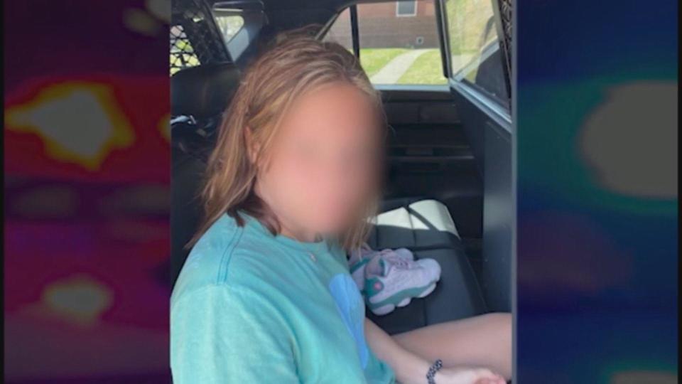The young girl is sitting in the back seat of the police car staring into the camera. She is wearing a green, short sleeved t-shirt and shorts. Her shoes are on the seat next to her. She has no make-up on and appears very young, dazed, and almost unrecognizable.  