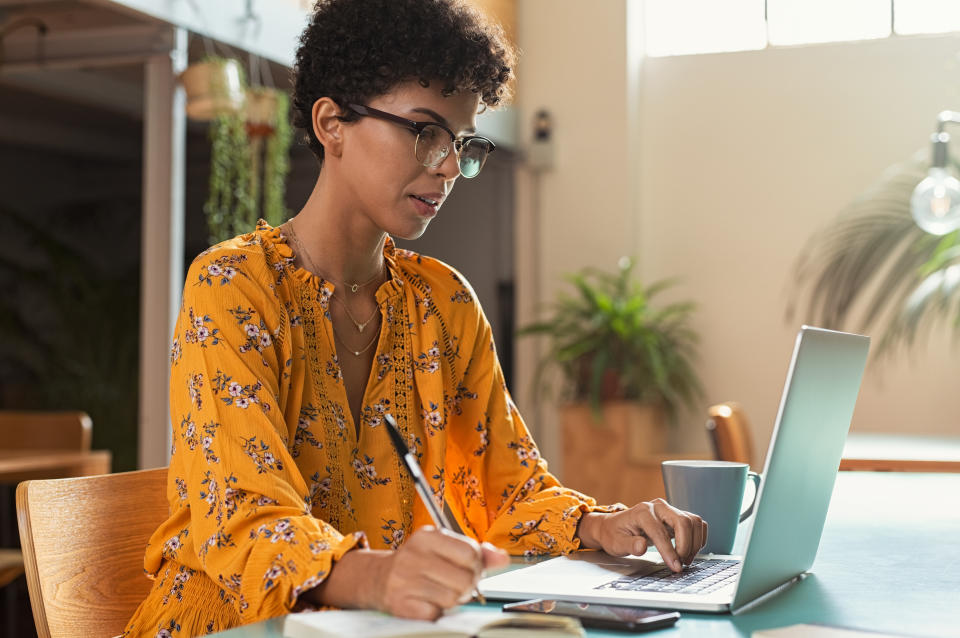 Focused college student sitting in cafeteria taking notes while using laptop. Young brazilian woman doing research for business at coffee shop. African american girl sitting in cafe using computer and writing notes."r