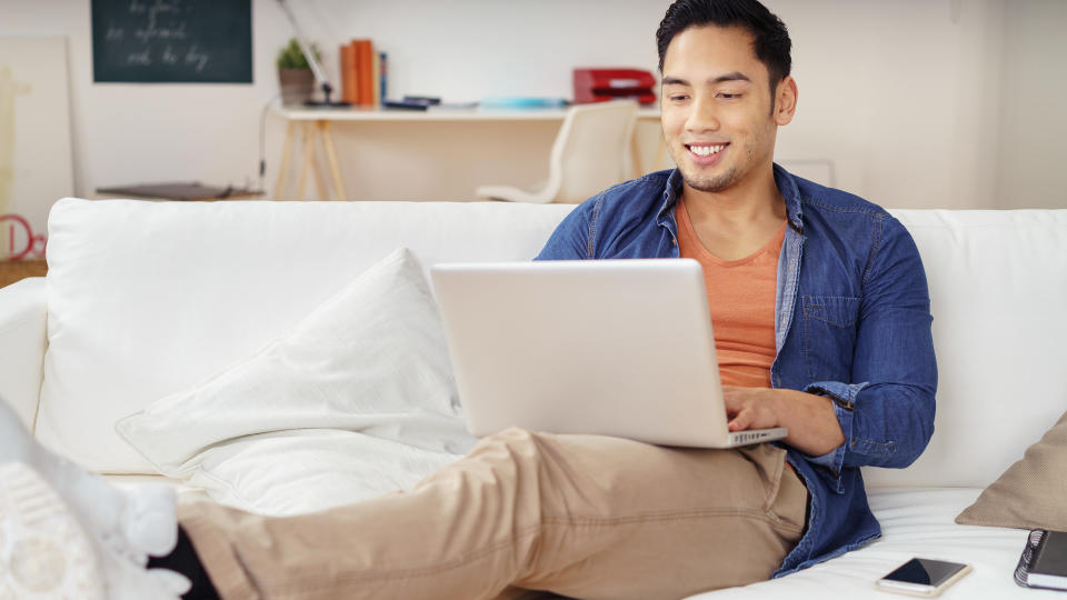Young Asian man spending a relaxing day at home with his feet up on a sofa surfing the internet on a laptop computer with a smile, fees, America, money, payment, avoid fees, bills, debt