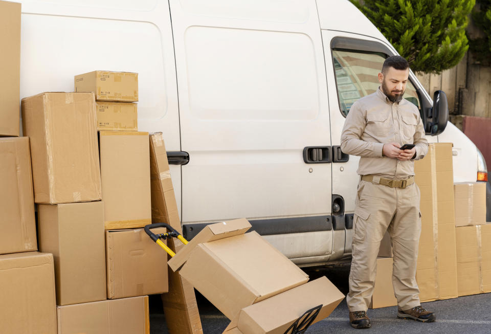 Delivery person with clipboard by a van with parcels, one package fallen over