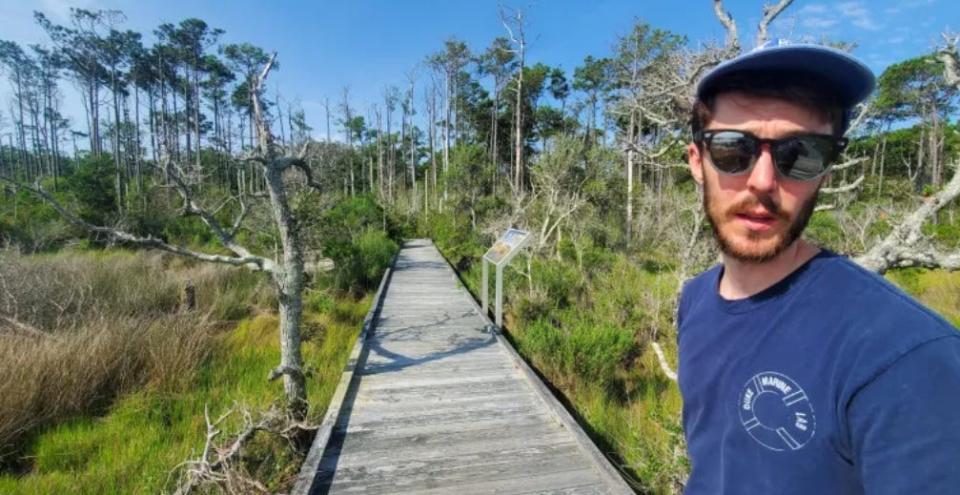 Patrick Gray, a doctoral candidate in marine science at the Duke University Marine Lab in Beaufort, N.C., in the foreground. Behind him are dead pines and oaks along the boardwalk of a nature trail in the Core Sounds National Seashore on Harkers Island. These trees perished after a tidal surge from Hurricane Florence overwhelmed the Down East peninsula.