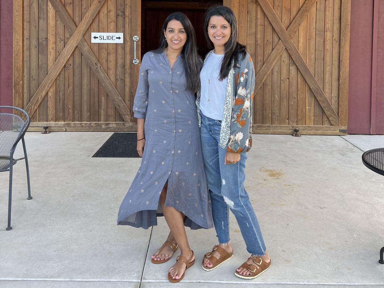 A photo of Neerja Patel on the left and Shailja Ambrose on the right. Neerja is wearing a flowy blue long-sleeve dress with a floral pattern and Shailja is wearing a floral jacket with yellow flowers, a light blue t-shirt, and blue denim jeans with a patched knee. They stand outside on a concrete floor in front of wooden sliding barn doors.