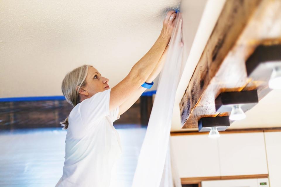 A mature woman is sticking blue painter's tape to the top of a drywall ceiling beam as part of preparing to paint her kitchen.