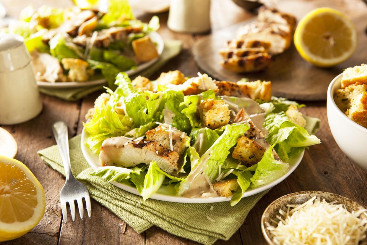 Chicken caesar salad on a white plate with a fork on a wooden table with another plate of chicken caesar salad and ingredients in the background