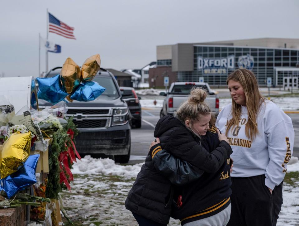 Girls hug in the snow in front of Oxford High School beside a memorial with balloons and flowers