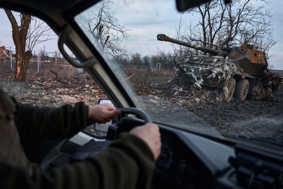 A view from inside a vehicle with hands on a steering wheel shows a damaged armored vehicle