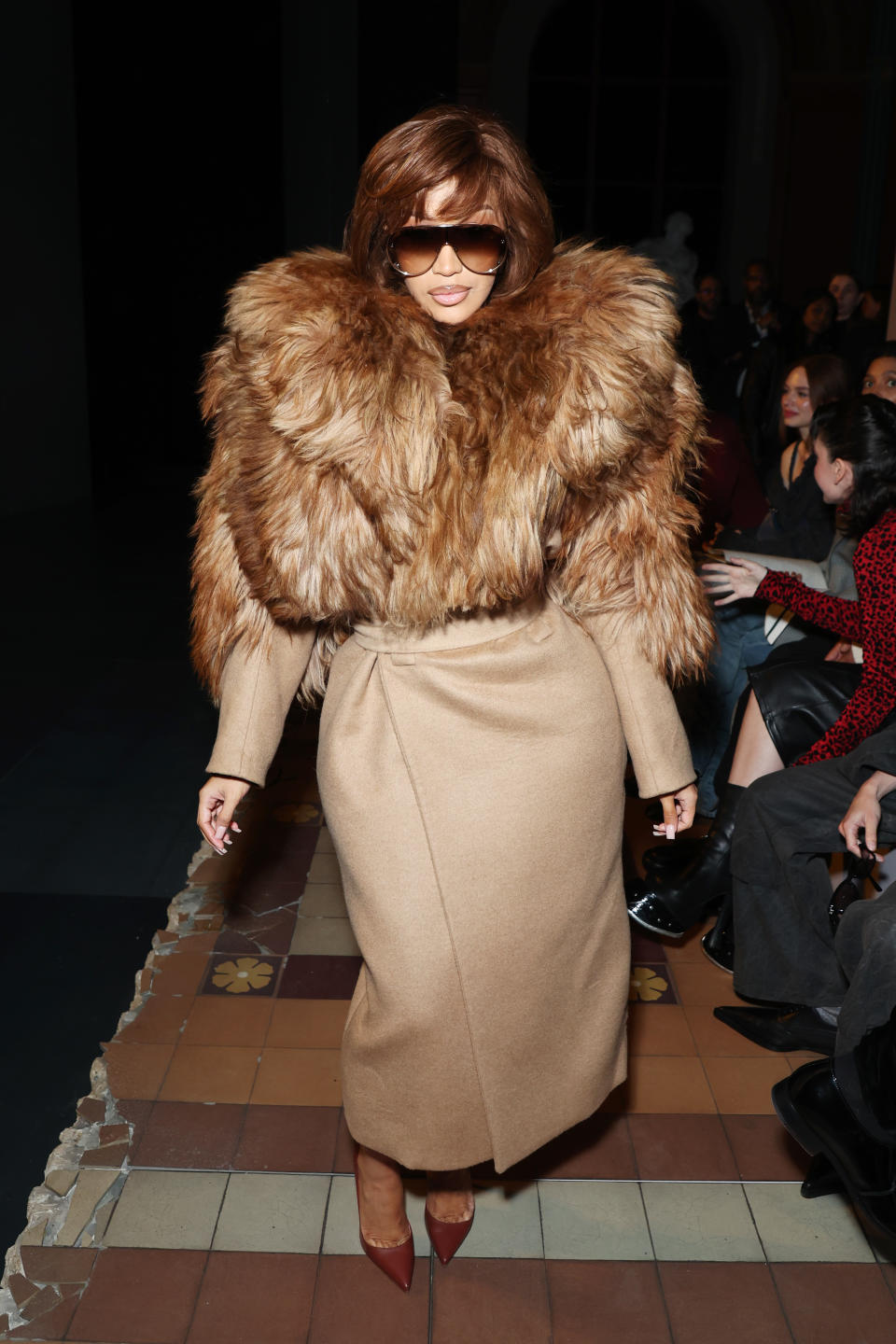 Cardi B stands confidently in an outfit with a large, fur-like collar and a long, belted coat and sunglasses