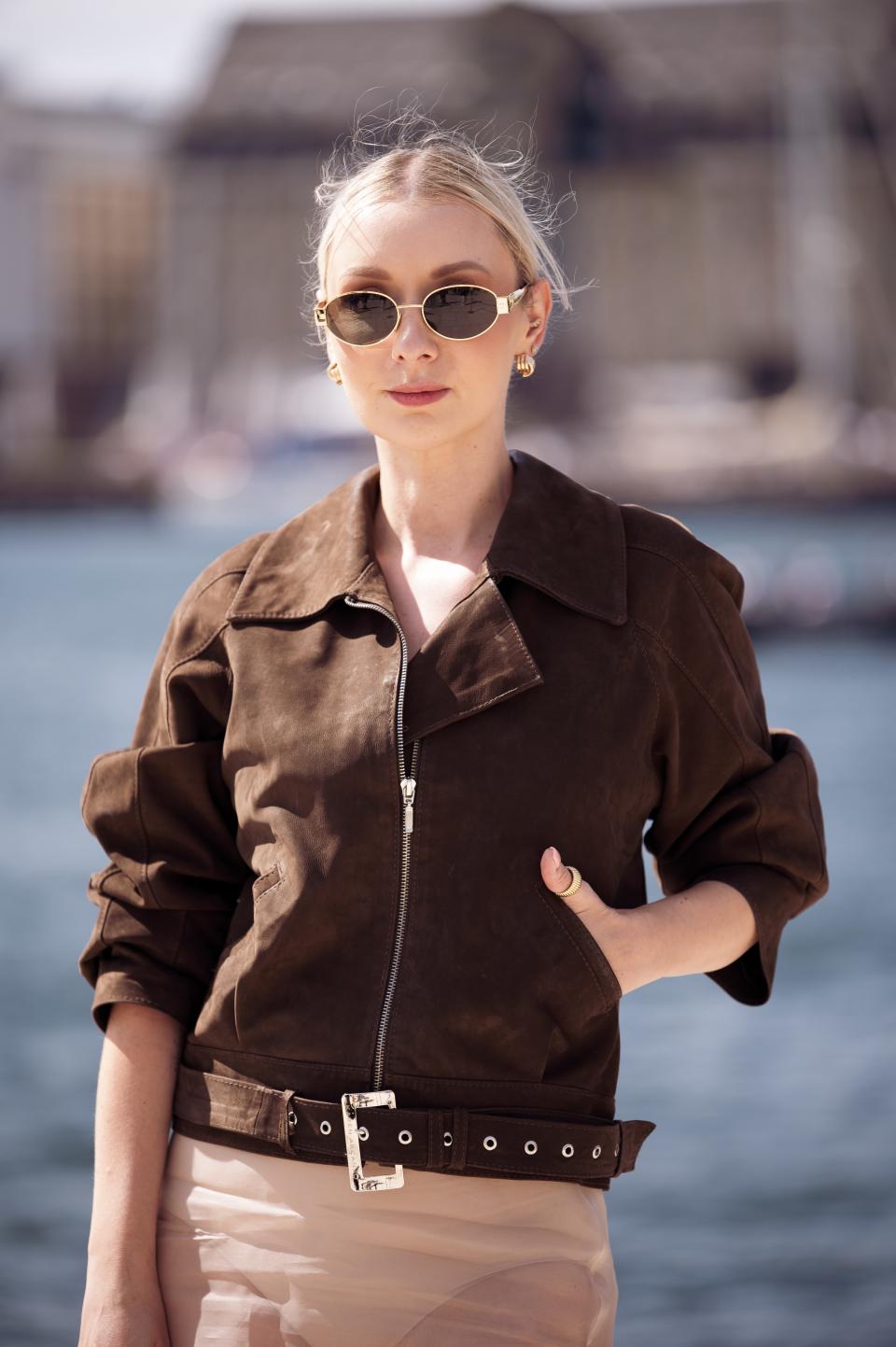 A woman in a stylish brown jacket with a belted waist and round sunglasses is standing outdoors near the water