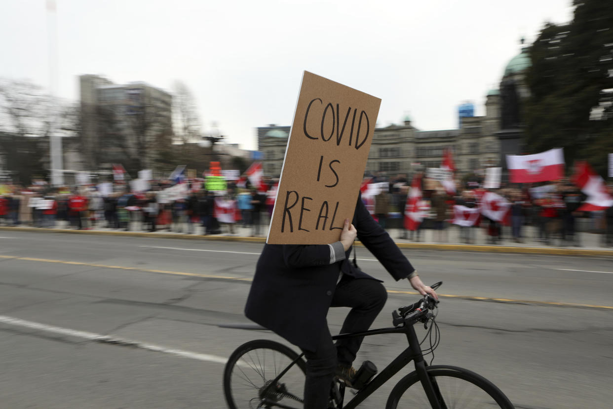 A counter-protester holds a sign while biking past supporters as they gather near the legislature to protest during a demonstration against COVID-19 restrictions in Victoria, British Columbia, Saturday, Feb. 5, 2022. (Chad Hipolito/The Canadian Press via AP)