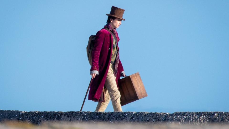 Timothée Chalamet como Willy Wonka, uma homem branco de casaco vinho, cachecol, bengala na mão e chapéu