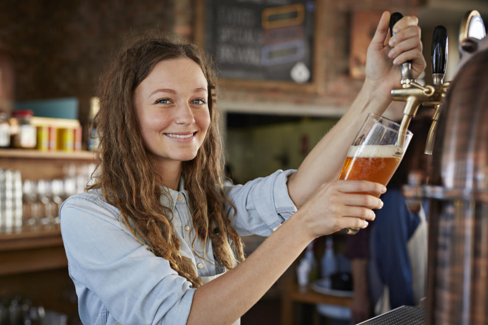 Smiling woman in casual clothing pouring a beer from the tap in a bar. In the background you can see the bar interior and people chatting