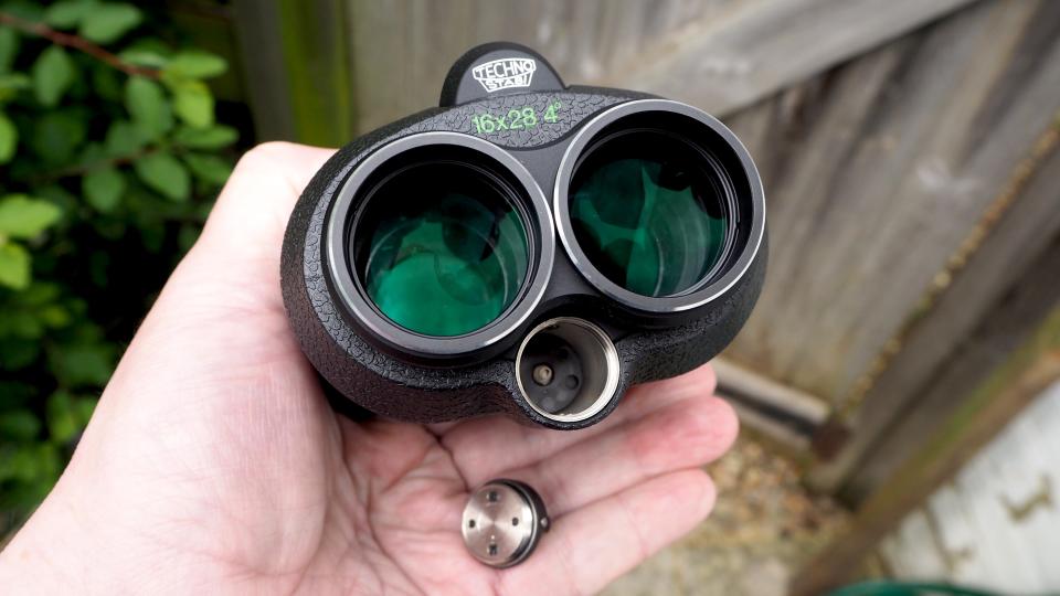Fujifilm Fujinon Techno-Stabi TS16x28WP binocular on held in a hand in front of a brown wooden fence
