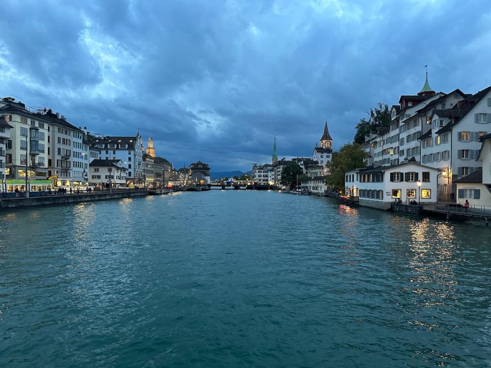 wide river lined with old zurich buildings with yellow lights on in the windows on a cloudy evening