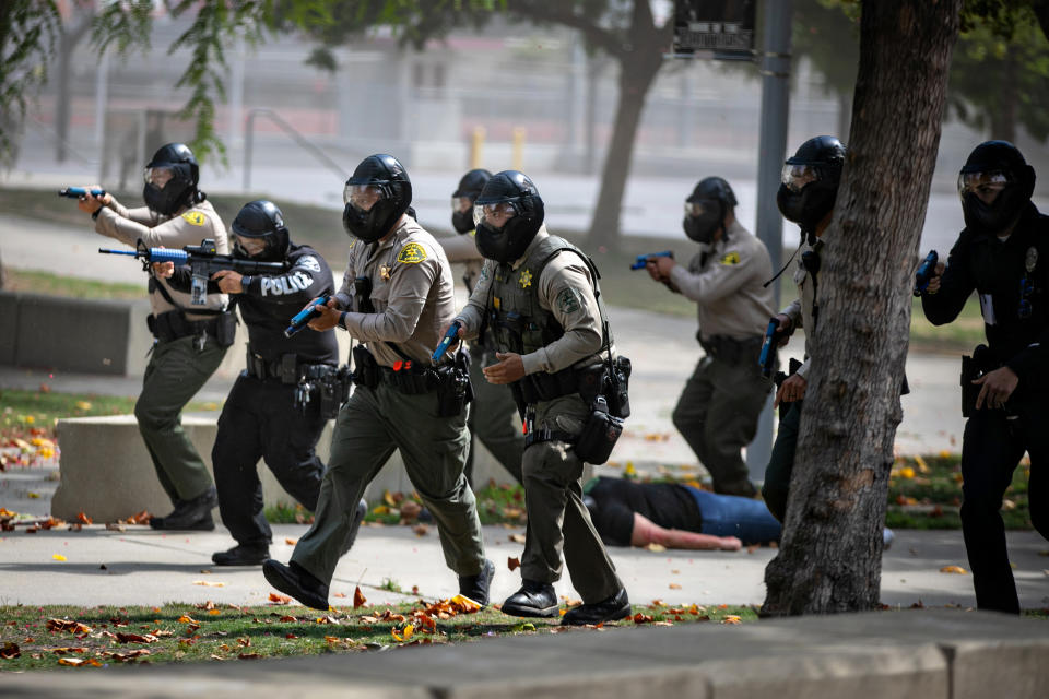 Eight sheriff's deputies and police officers in helmets march down a sidewalk, guns at the ready.