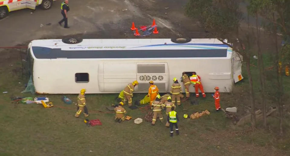 The bus can be seen on its left side on grass with first responders gathered around the vehicle. 