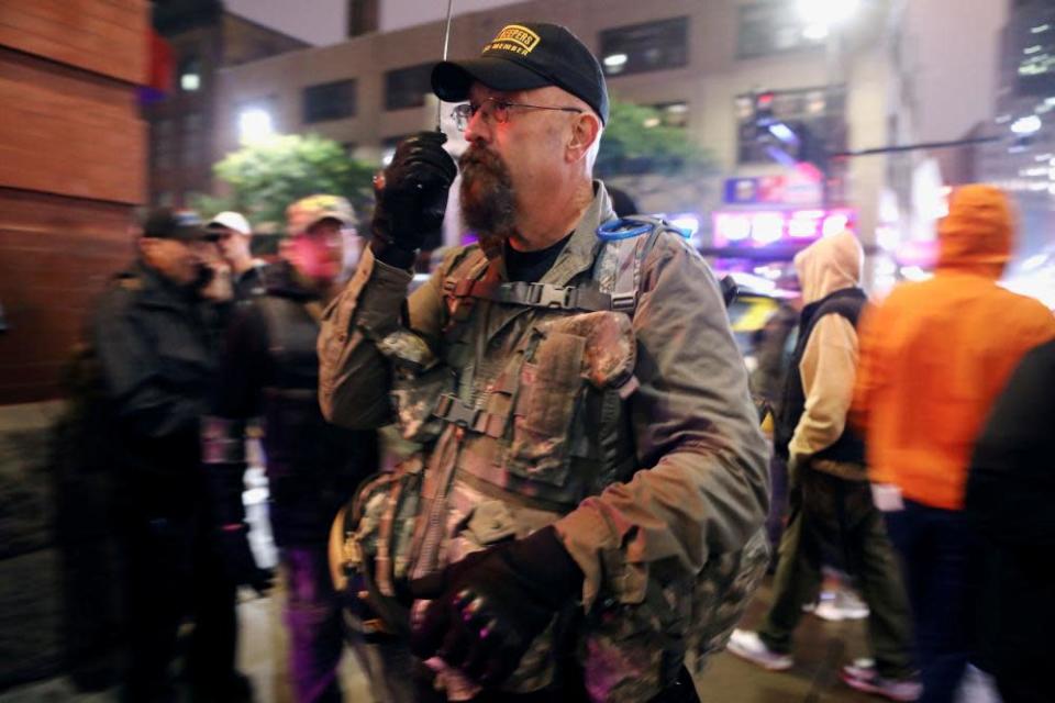 A man wearing military garb and a black baseball cap speaks into a radio as he monitors a crowd.