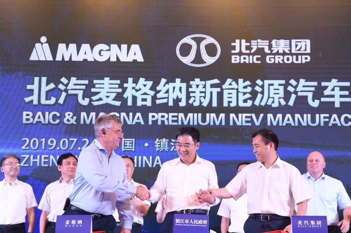 Magna and BAIC executives are shown shaking hands in front of a banner with the two companies' logos and text in both English and Chinese.