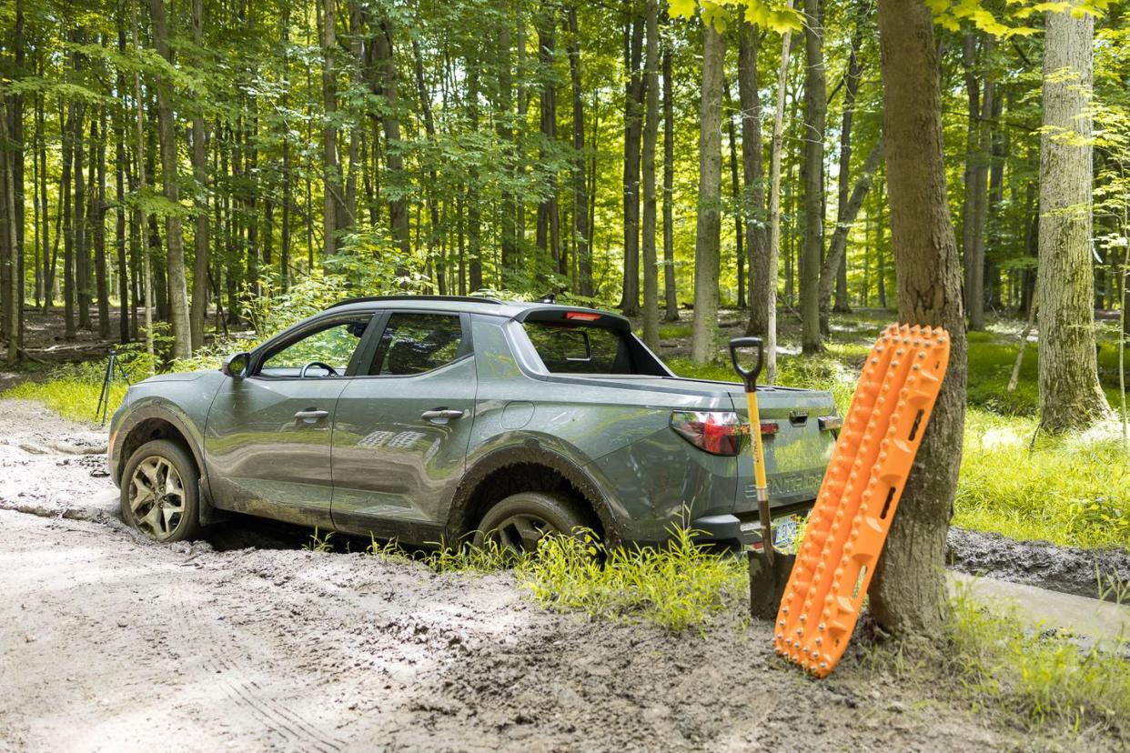 a car parked in a wooded area, stuck in mud, with a shovel and traction boards leaning against a tree in preparation for recovery