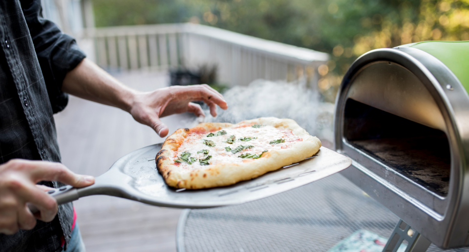 man holding pizza outside and green and silver outdoor pizza oven, pizza oven outside on balcony in backyard