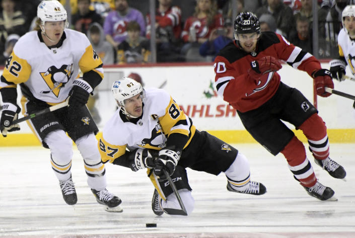 Pittsburgh Penguins center Sidney Crosby (87) reaches for the puck as New Jersey Devils center Michael McLeod (20) and Penguins right wing Kasperi Kapanen (42) look on during the first period of an NHL hockey game, Sunday, Dec.19, 2021, in Newark, N.J. (AP Photo/Bill Kostroun)