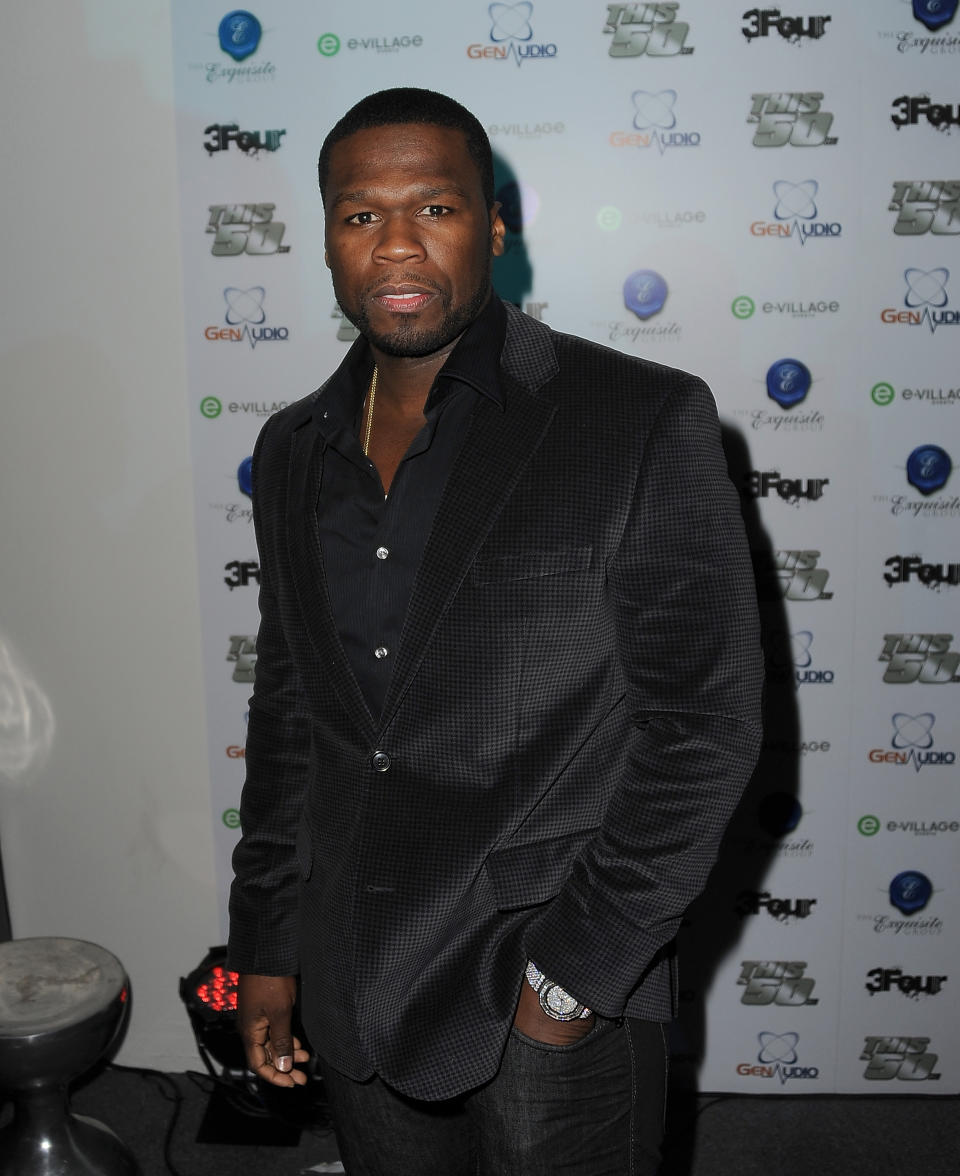 50 Cent en costume noir et chemise pose devant une toile de fond de marque lors d'un événement