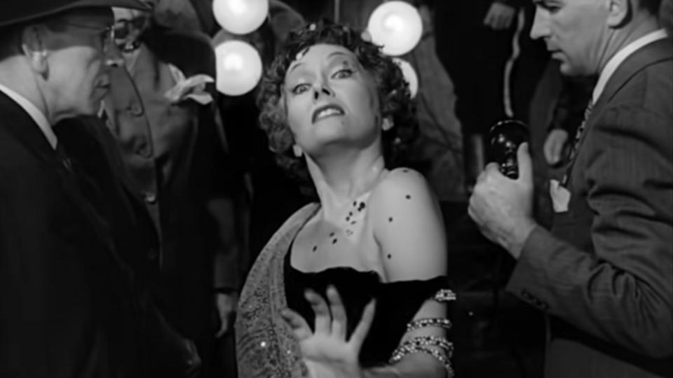 Gloria Swanson, in makeup and dress, walking in front of police cameras in Sunset Boulevard