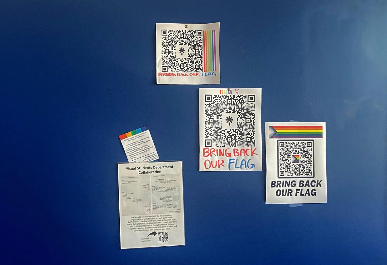 Only posters and thumbtacks hang on a wall at the back of a cafe where a pride flag used to be at St. Edward's University. The posters have a QR code with the message: "BRING BACK OUR FLAG."