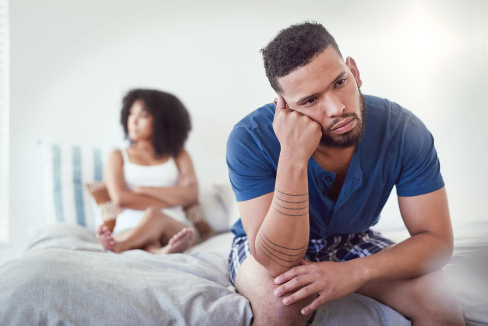 A man and a woman sitting separately on a bed, looking away from each other, appearing upset