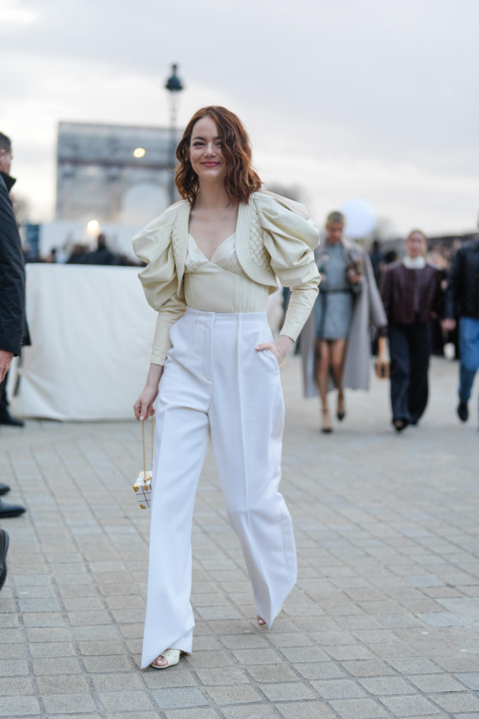 Emma Stone is wearing a blouse and a jacket with voluminous sleeves along with high-waisted trousers. She is holding a small box purse and smiling while walking outdoors to an event