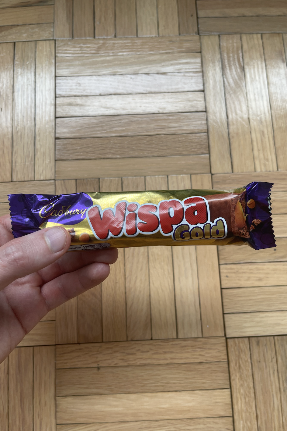 Hand holding a Cadbury Wispa Gold chocolate bar over wooden floor