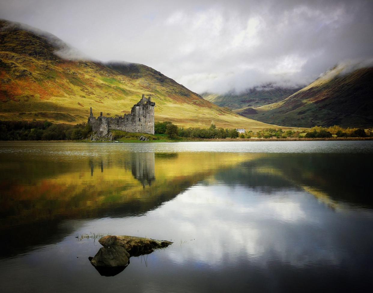 ancient castle amid rolling, treeless scottish hills with clouds and mist with a small lake reflecting sky in front of it