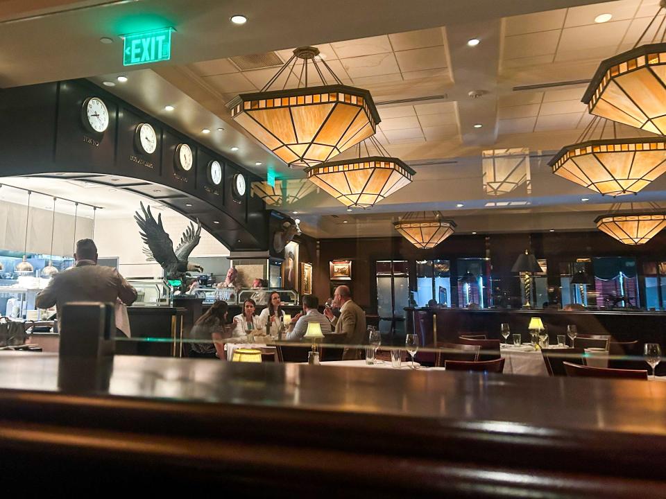 The interior of a Capital Grille restaurant, with tables covered with white tablecloths, chairs, chic chandeliers, and a view into the kitchen.