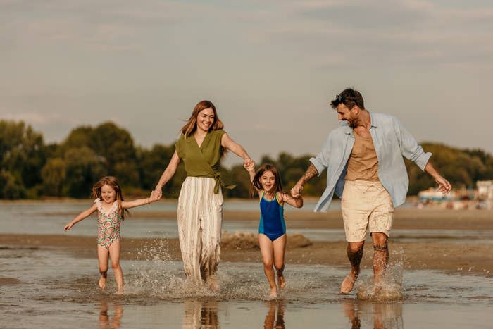 A family of four, consisting of a man, woman, and two young girls, walk hand-in-hand along a beach, smiling and enjoying the water