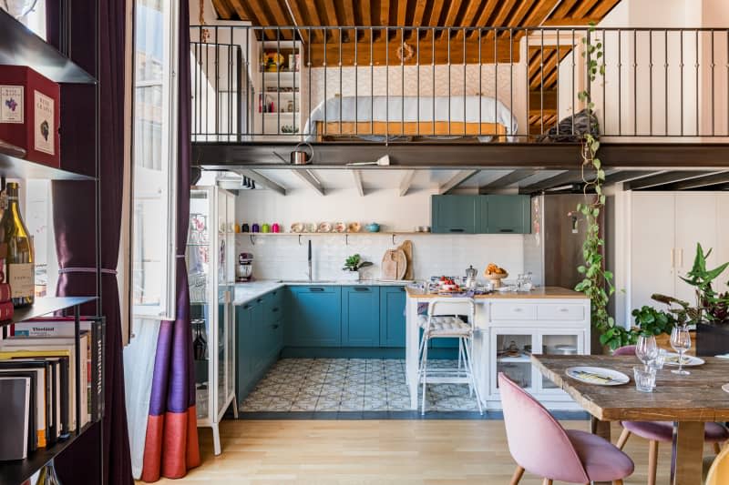 A kitchen with white tiled backsplash and teal cabinets below a lofted area.