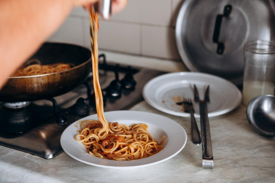 A person is serving spaghetti from a frying pan into a white plate, with utensils and additional cookware visible on the stove and countertop