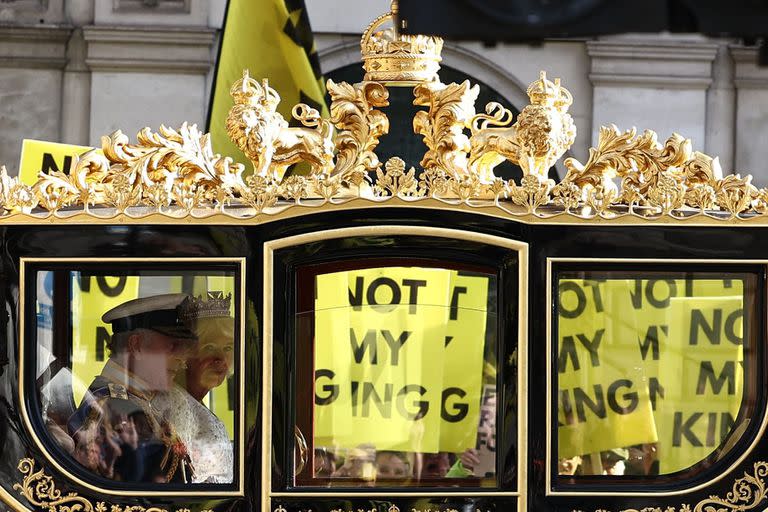 El rey Carlos III de Gran Bretaña y la reina Camilla de Gran Bretaña viajan en carruaje mientras manifestantes portan pancartas que dicen 