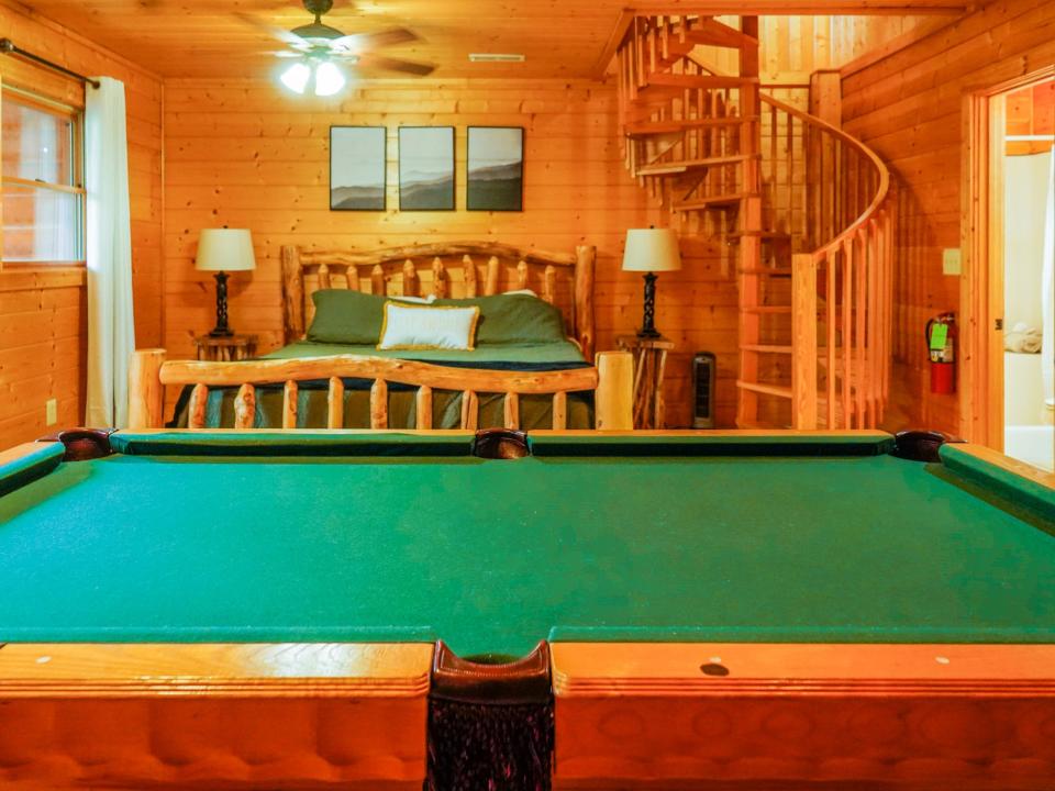 A wide view of the bedroom shows a pool table with green felt in front of the bed with a green spread on the left with nightstands holding lamps on either side. There's a celing fan above the bed and the spiral staircase is on the right.