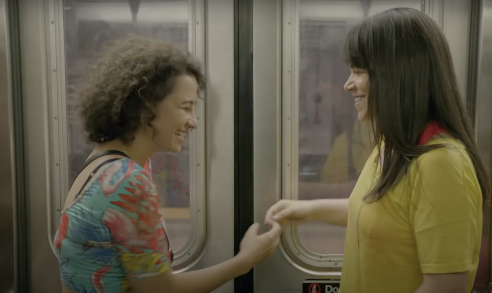 Ilana Glazer and Abbi Jacobson smile at each other while holding hands on a subway train