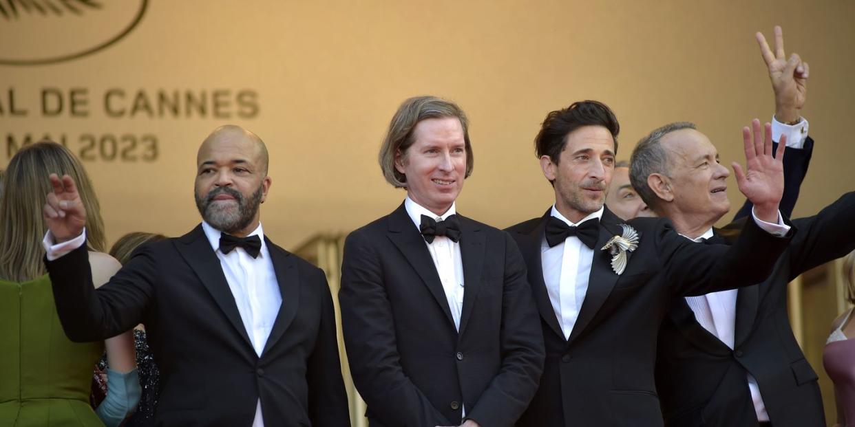 jeffrey wright, wes anderson, adrien brody, and tom hanks alls tand on a stage waiving at people, wearing tuxedos