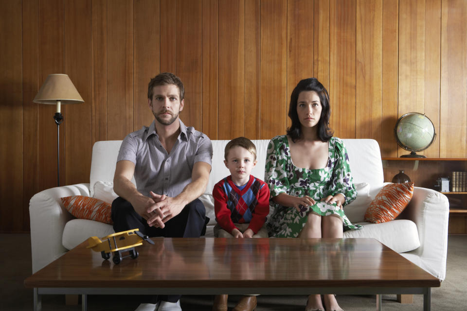 Two adults and a child sit on a couch with a toy truck on the table in a home setting