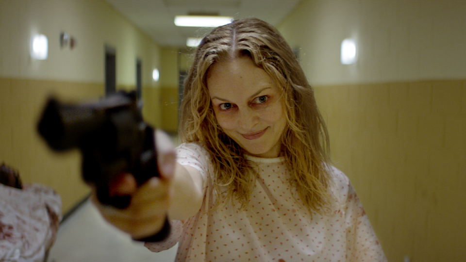Heather Graham wears a hospital gown and grins while pointing a gun in the Lovecraft adaptation Suitable Flesh.