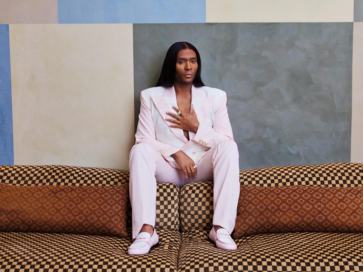 Law Roach sitting on a couch, pink suit, long straight hair