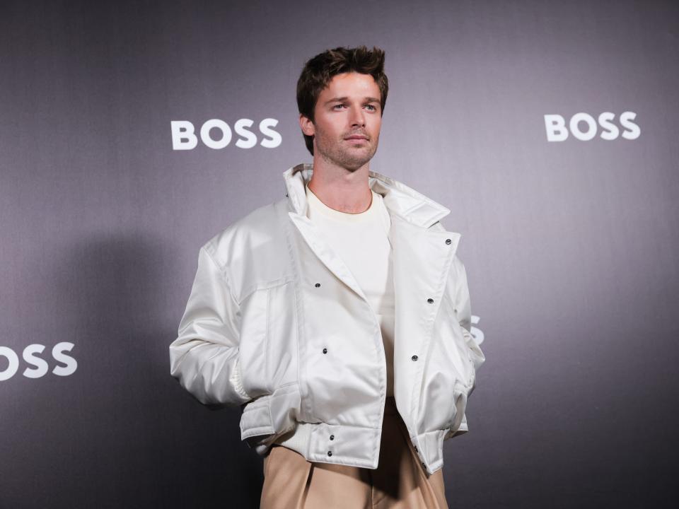 Patrick Arnold Schwarzenegger wearing an oversized silver jacket and chino pants while standing in front of a black wall with the logo for Boss