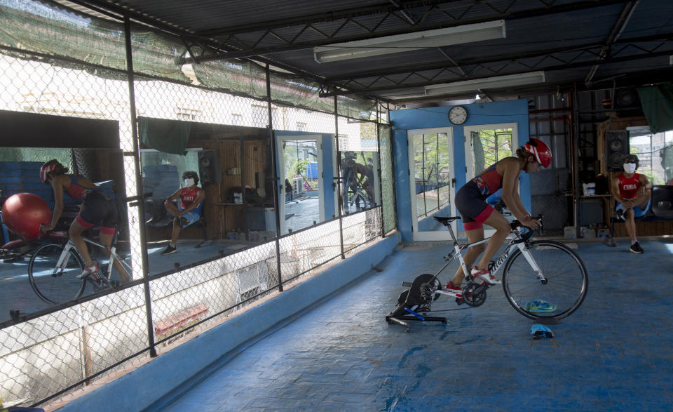 Cuban triathlon athlete Leslie Amat rides her bicycle, adapted to be stationary, under the watch of her coach Dioseles Fernandez in the patio of her home in Havana, Cuba, Monday, April 20, 2020. Amat aims to classify for the 2021 Tokyo Summer Olympics, using improvised equipment helping her to train amid a quarantine triggered by the spread of the new coronavirus. (AP Photo/Ismael Francisco)