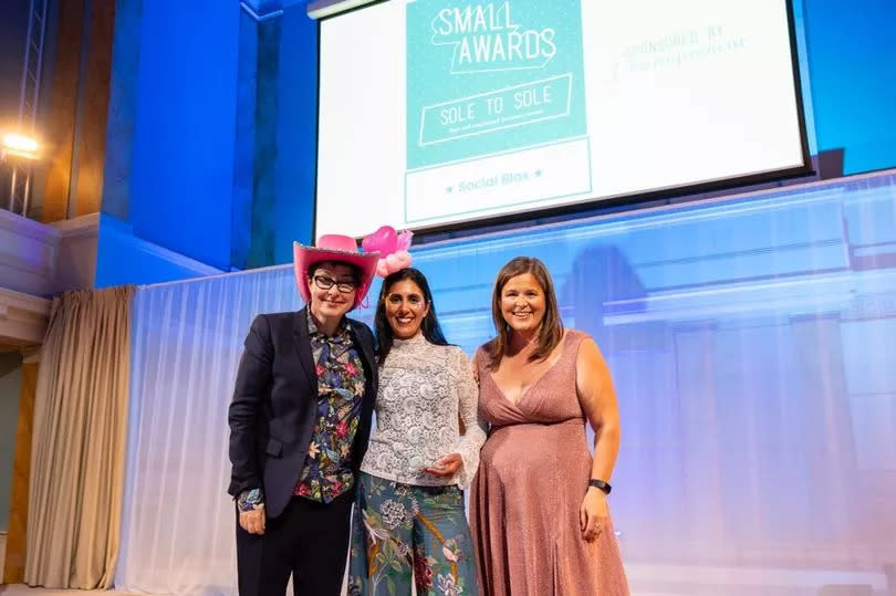 A business owner is presented an award on stage, three women smile for the camera including comedian Sue Perkins