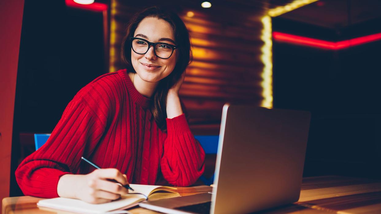 Smiling young woman enjoying freelance work in cafe interior making notes of ideas in notepad, positive female student doing homework in good mood satisfied with wifi for learning on laptop computer - Image.